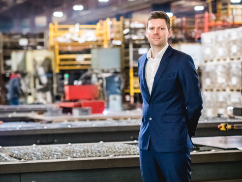 Commercial Director of BM Catalysts Mark Blinston is stood next to heavy machinery. He is dressed smart and is smiling.