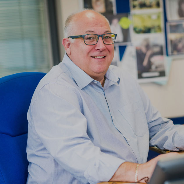 A customer support team member is sat in an office chair. He is smiling and wears a smart shirt and glasses.