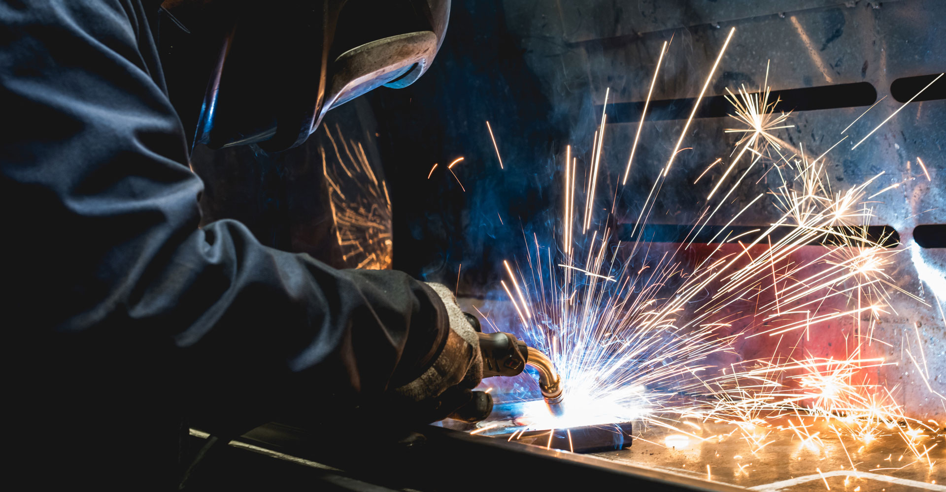 A man is welding in a factory. Sparks are flying and he is wear overalls and a mask.