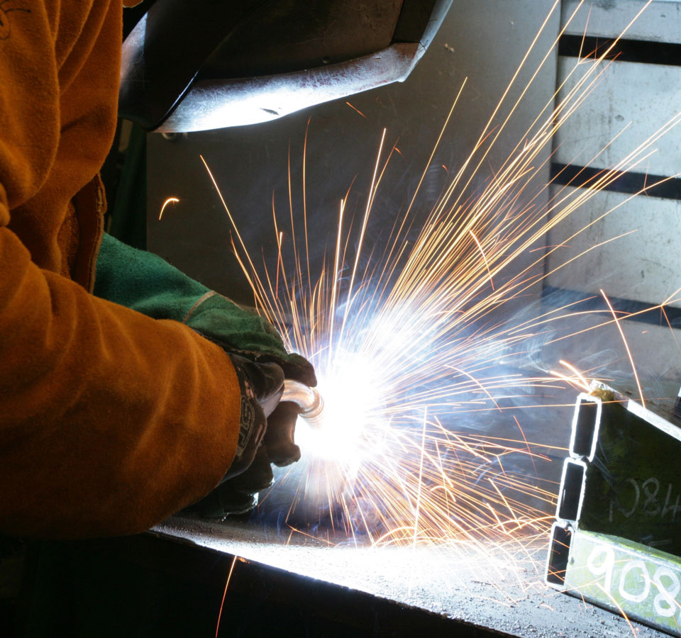 A man is welding as sparks are flying and he is wear overalls and a mask.
