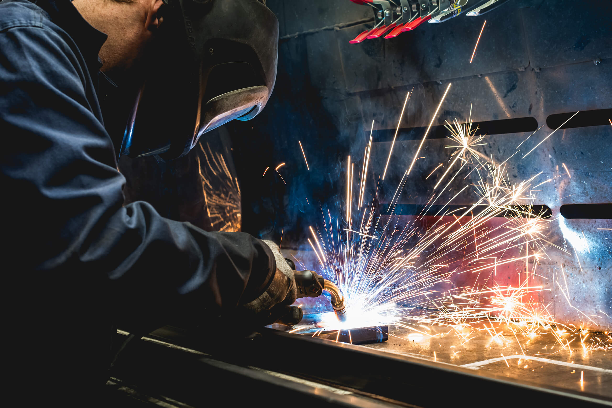 A man is welding in a factory. Sparks are flying and he is wear overalls and a mask.