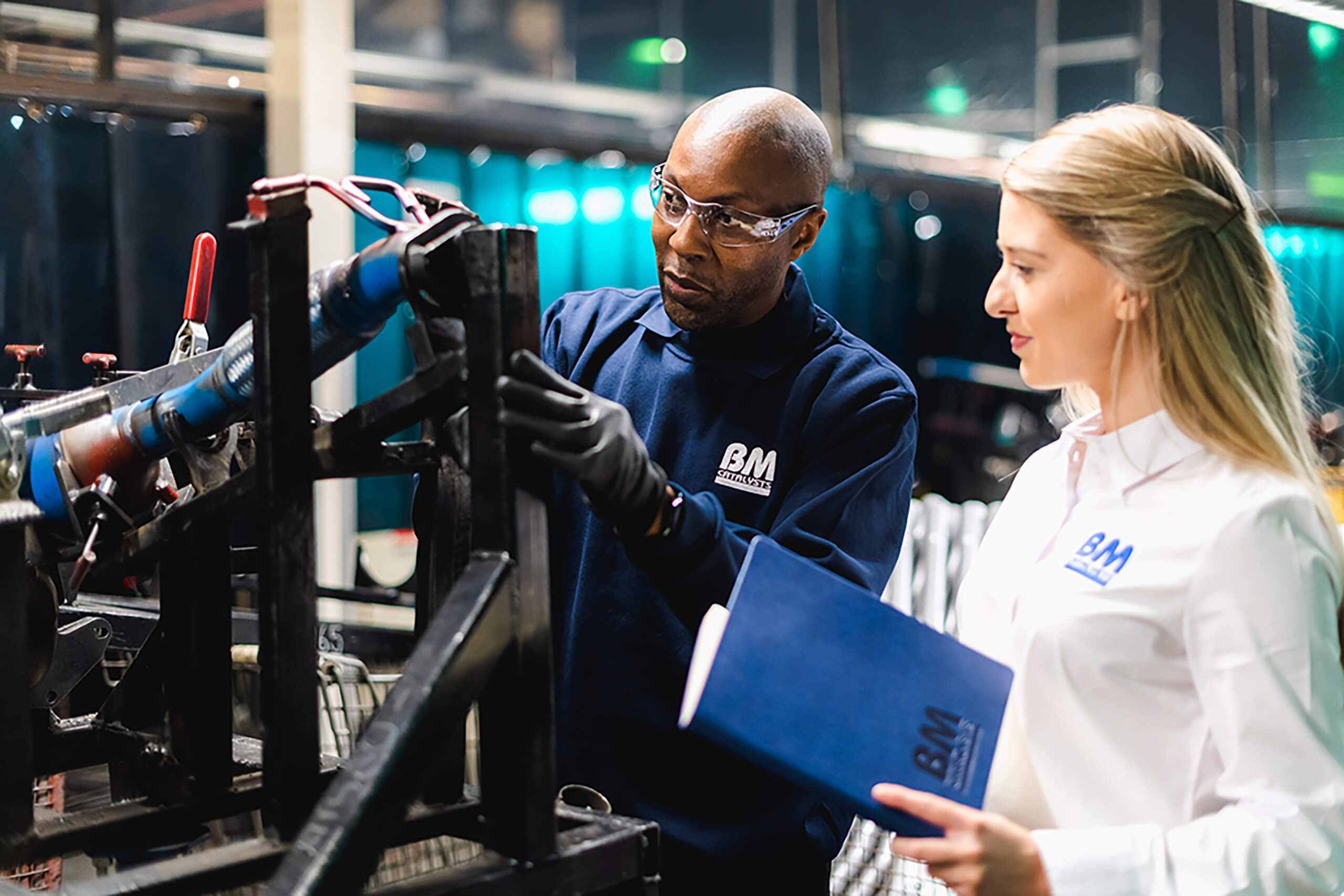 A manufacturer is working on a product part in the factory as his colleague is stood next to him observing. The manufacturer is wearing google and his colleague has a note book.