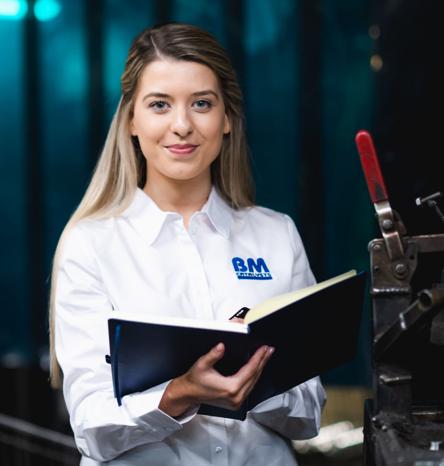 A lady is smiling as she stands next to machinery in a manufacturing facility. She is in a smart shirt and is holding a A4 note book which is open.