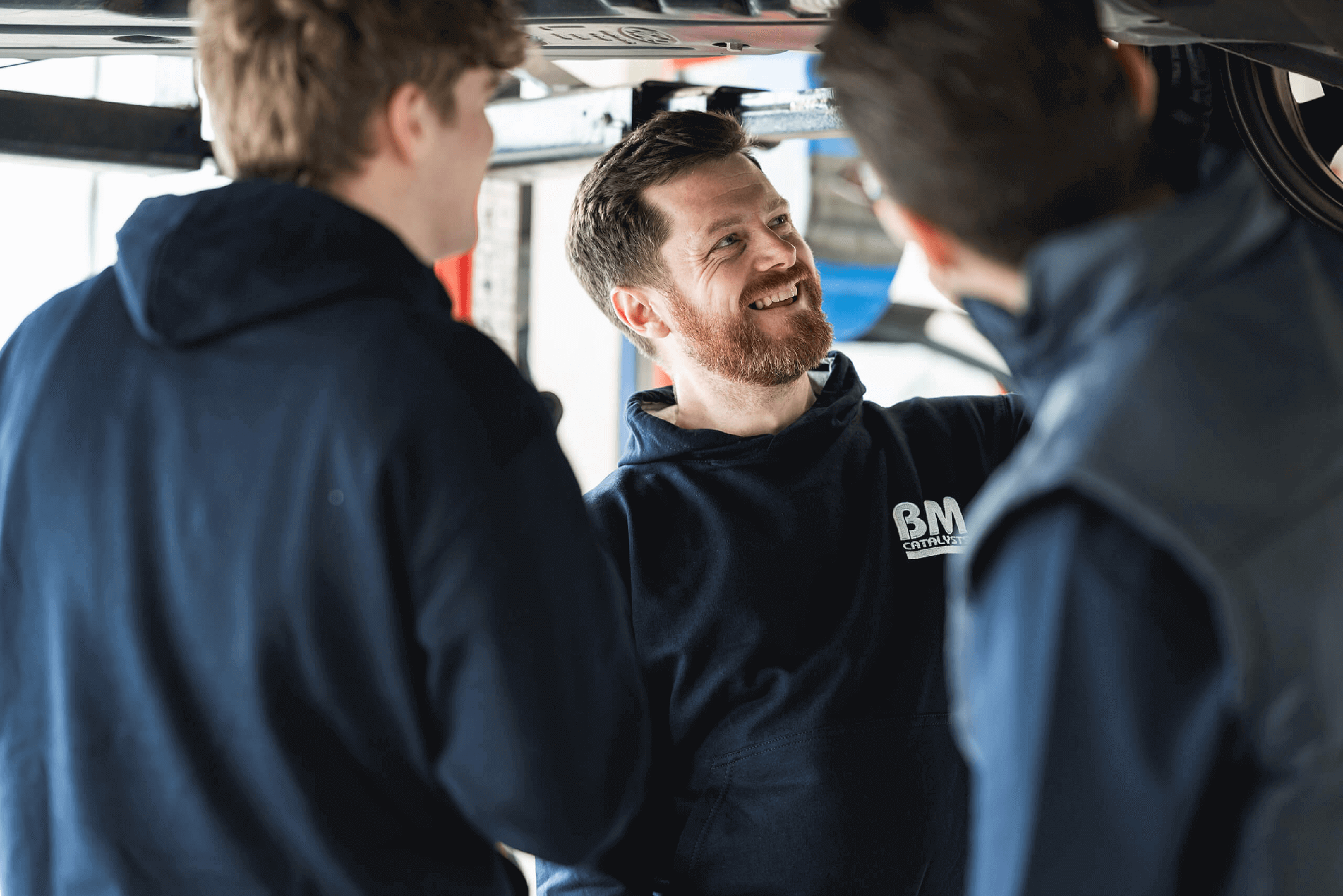 3 garage fitters are stood underneath a lifted car in a garage workshop.