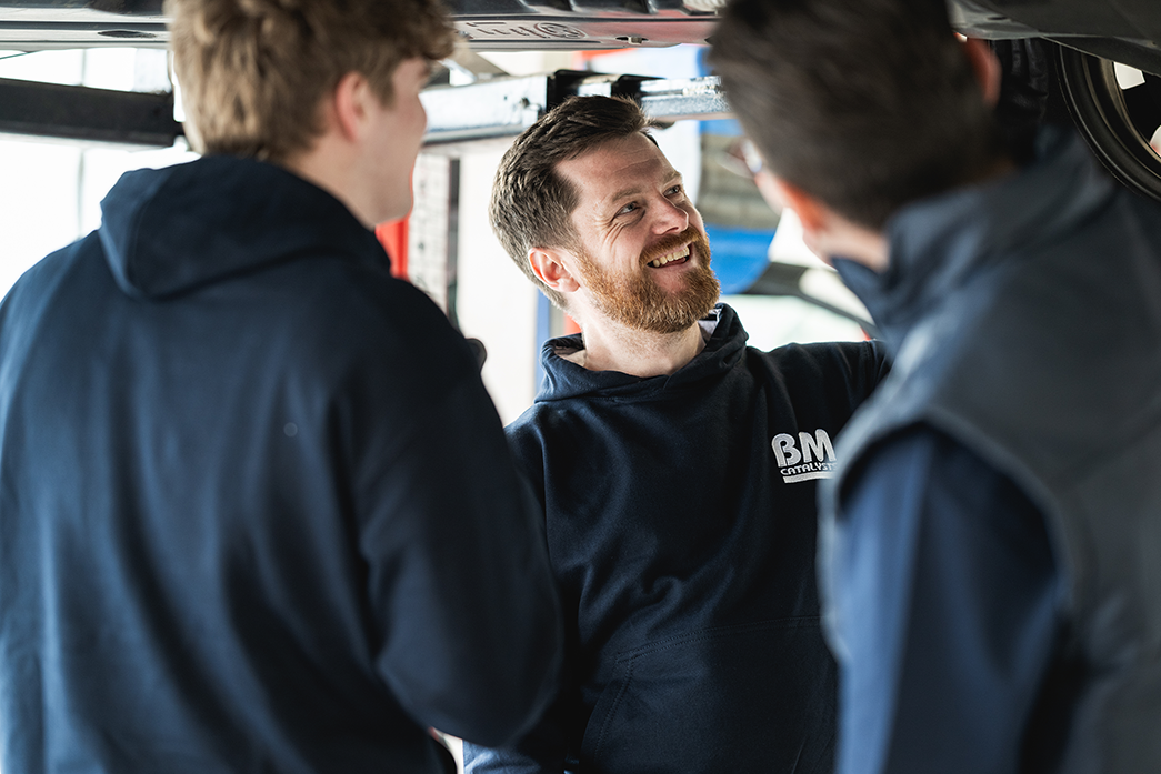 3 garage fitters are stood underneath a lifted car in a garage workshop.