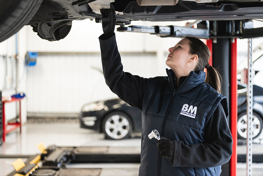 A woman is checking underneath a car in a garage workshop. The car has been lifted above her.